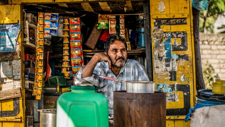 A man is sitting in his tea shop with a forlorn expression. A huge iron tea canister, green plastic bucket and other wares are seen in the surrounding.